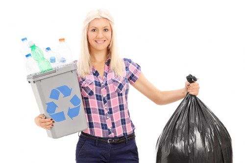 A young woman with long blonde hair, dressed in a short-sleeved, pink and blue checkered shirt and dark trousers, standing against a plain white background, holding an outdoor grey recycling bin filled with various plastic bottles and containers in her left hand, and a closed black rubbish bag in her right hand, exhibiting a neutral facial expression. The recycling bin features a blue recycling symbol on the front. The image appears to promote waste and rubbish removal services, with the clear focus on sorting recyclables and general waste collection, relevant to environmental initiatives in Marylebone and surrounding areas.