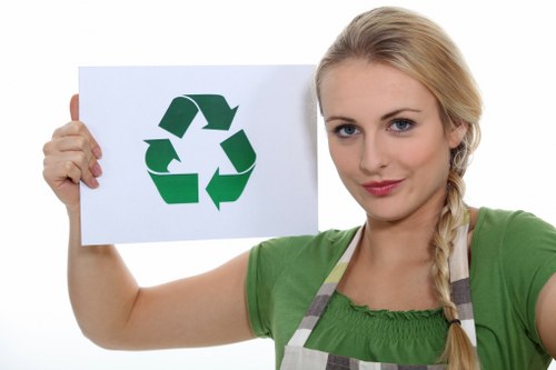 A young woman with blonde hair styled in a side braid, wearing a green short-sleeved shirt and a light grey and white striped apron, holds up a white sheet of paper featuring a large green recycling symbol composed of three arrows forming a triangle. She is standing against a plain white background. The image emphasizes environmental awareness and recycling, reflecting areas served by Office Clearance Marylebone in London, and aligns with rubbish removal services focused on sustainable waste management in the Marylebone area.