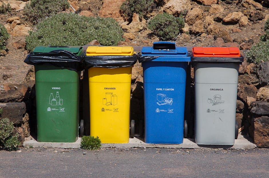 Four separate waste collection bins lined up along a paved surface in an outdoor setting, positioned against a background of rocks and sparse shrubbery. The first bin from the left is green with a bla