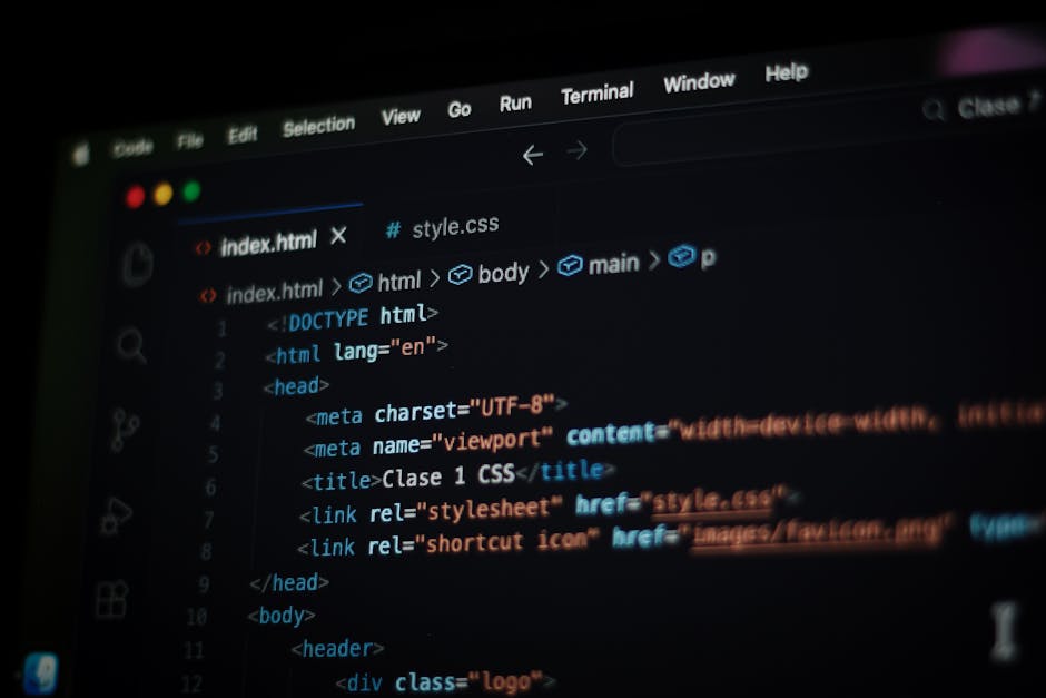 A person with short dark hair, dressed in a light blue shirt, working on a laptop at a desk. The laptop screen displays lines of multicolored programming code, primarily in red, blue, yellow, and whit